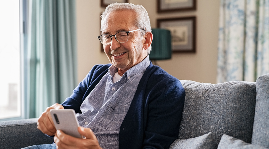 Ein grauhaariger Mann in Jeans und mit Brille sitzt auf einem Sofa. Er schaut in sein Smartphone und lächelt.