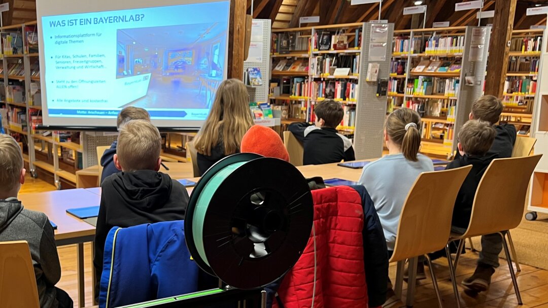 In der Stadtbücherei Mühldorf am Inn sitzen ca. 10jährige Kinder an einem Tisch und schauen auf eine Leinwand, auf der das BayernLab sich vorstellt. Im Hintergrund sind hohe Regale mit Büchern.