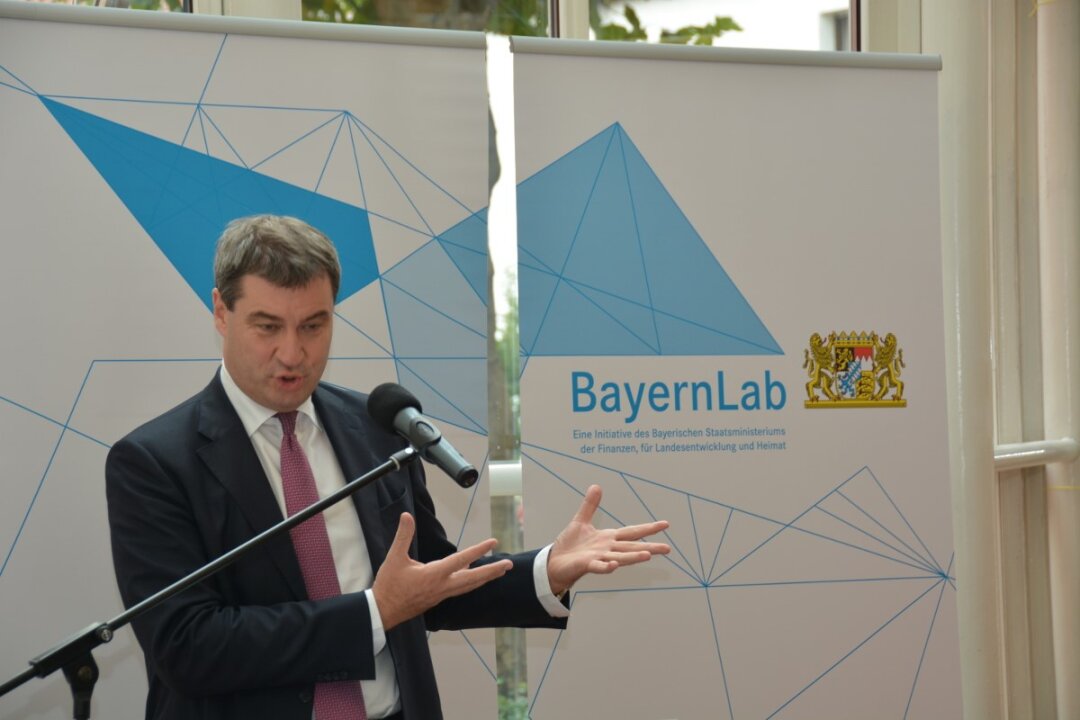 Finanz- und Heimatminister Söder trägt ein dunkelblaues Jacket, weißes Hemd und Schlips. Er spricht in ein Standmikrofon und hat beide Hände geöffnet. Im Hintergrund ist das Logo vom BayernLab zu sehen und die für die BayernLabs typischen blauen und weißen Polygone.