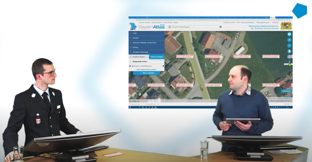 Im Vordergrund stehen zwei Männer hinter einem Tresen, die sich gegenseitig anschauen. Der linke Mann ist der Referent von der Feuerwehr. Er trägt eine Feuerwehr Uniform mit Sakko, Hemd und Krawatte. Der rechte Mann ist der Moderator vom BayernLab. Im Hintergrund des Moderators befindet sich ein Kartenausschnitt aus dem BayernAtlas mit roten Beschriftungen auf Häusern.