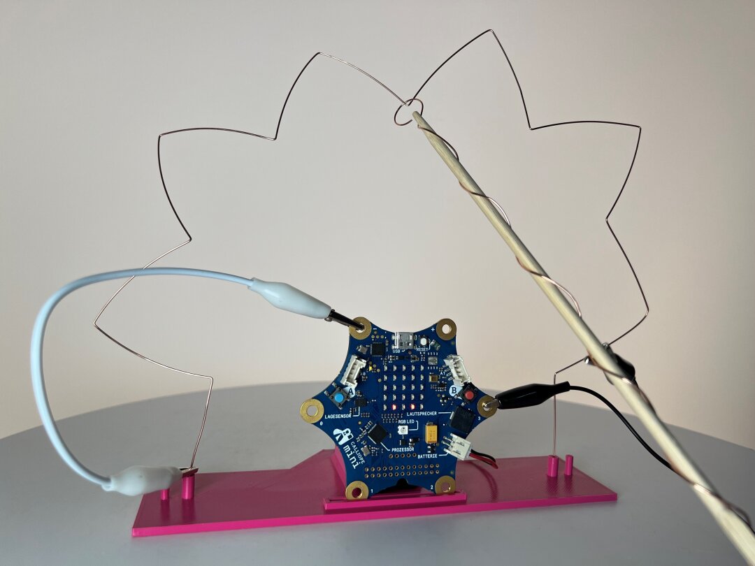 Eine sternförmigen Platine steckt aufrecht in einer rosafarbenen Halterung. An ihr sind zwei Krokodilklemmen befestigt. Ein mit Draht umwickelter Holzstab hat eine Schlinge am vorderen Ende und wird um einen zackenförmig gebogenen Draht geführt. 